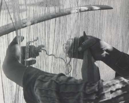 Close-up view of a person’s hands weaving threads on a loom, with light filtering through the vertical strands.