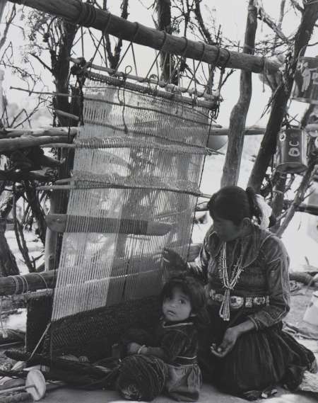 A woman weaves at a loom outdoors while a young child sits beside her, both dressed in traditional clothing. Branches and simple wooden beams frame the weaving area.