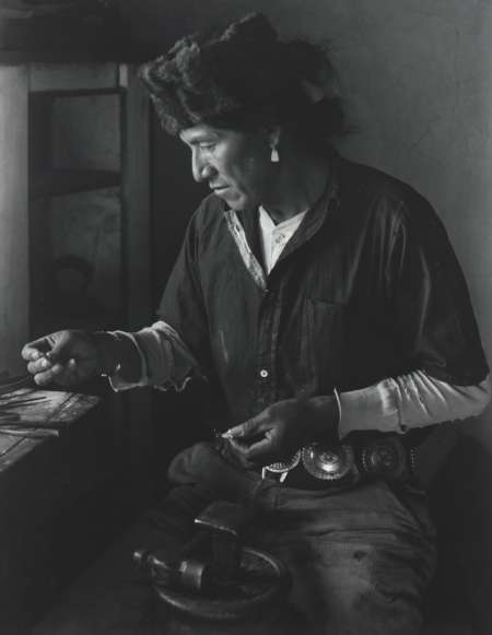 A person wearing a fur hat and patterned belt examines tools or objects at a workbench, lit from the side in a dim room.