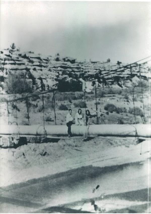 Three people sit on a large pipe bridge suspended over a dry, rocky landscape with shrubs and cliffs in the background.