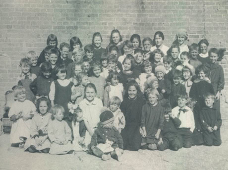 A large group of children pose for a black-and-white photo in front of a brick wall, most of them smiling.