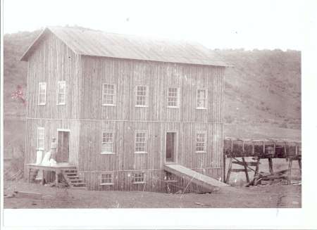 A two-story wooden building with multiple windows, a sloped ramp, and a raised walkway on the right, set against a hilly landscape.