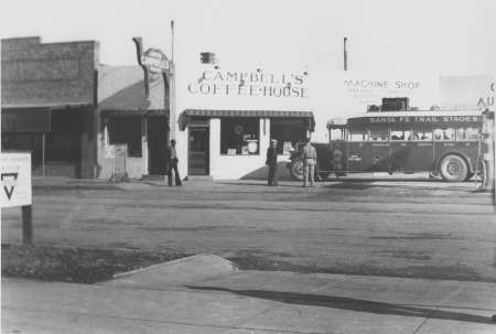 Black and white photo of Campbell’s Coffee House next to a machine shop, with a parked bus labeled "Santa Fe Trail Stages" and several people standing nearby.