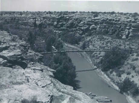 Black and white photo of a narrow pedestrian bridge spanning a deep rocky canyon with a river flowing below and sparse vegetation on both sides.