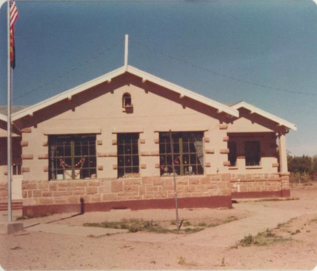 A single-story building with large windows and a stone foundation, under a clear sky, with a flagpole on the left and a concrete walkway in front.