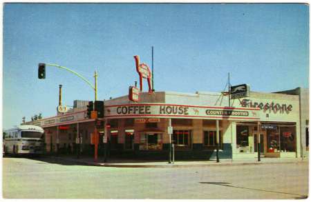 A street corner diner with large "Coffee House" and "Firestone" signs, a bus parked nearby, and vintage cars parked along the curb under a clear sky.