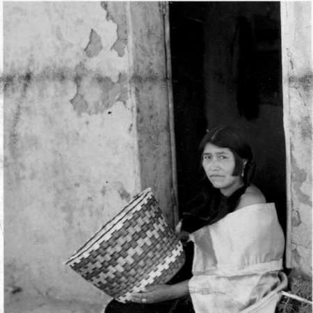 A woman sits in a doorway holding a large woven basket, against a textured, worn wall.