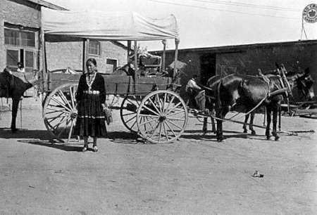 A woman stands in front of a covered wagon hitched to two donkeys or mules in a dusty outdoor setting, with buildings visible in the background.