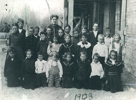 Black and white photo of a group of schoolchildren and their teacher posing outside a brick building. "1909" is written on the ground in front of them.