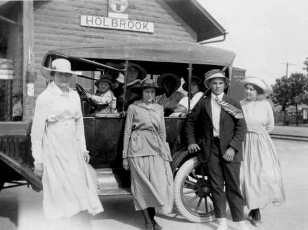 A group of people in early 20th-century clothing pose by a vintage car in front of a building with a "Holbrook" sign.