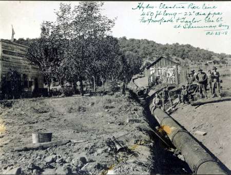 Historic black-and-white photo showing workers next to a large pipeline, with buildings, trees, and hills in the background; handwritten notes appear on the image.