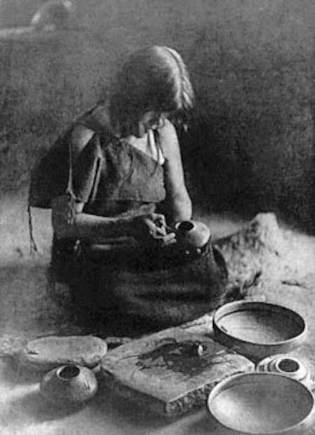 A woman sits on the ground, shaping pottery with her hands. Various clay vessels and tools are arranged around her. The setting appears rustic and traditional.