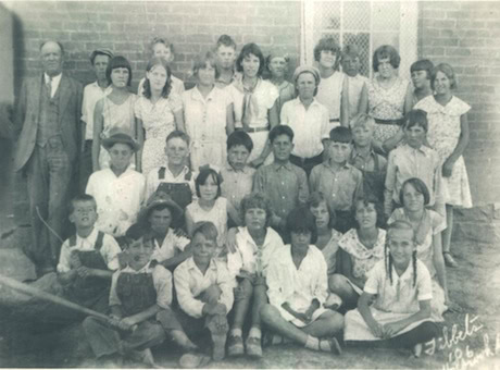 Black-and-white photo of a group of school children and an adult, posing in rows in front of a brick building, taken in 1926.