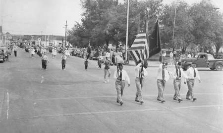 A group of people march down a street in a parade, with some carrying American flags; spectators and parked vehicles line the street.