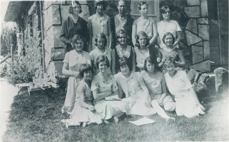A black and white photo of thirteen women and one man posing in two rows on grass beside a stone building. All are dressed in early 20th-century attire.