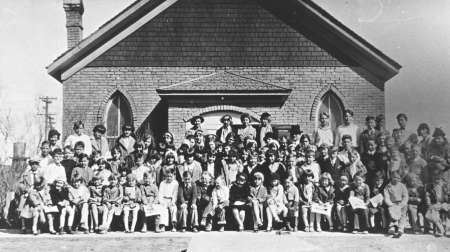 A large group of children and adults poses in front of a brick building with arched windows, likely a school or church, for a group photograph.