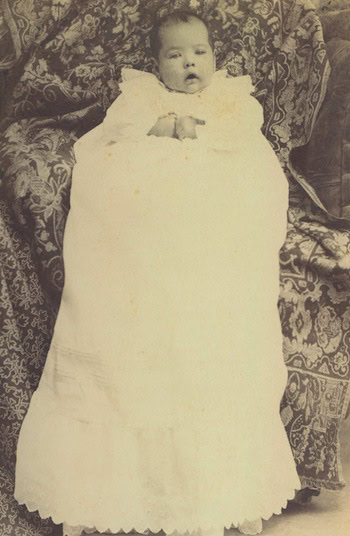 A vintage photograph of a baby lying on a patterned couch, wrapped in a long, white christening gown with lace and scalloped edges.