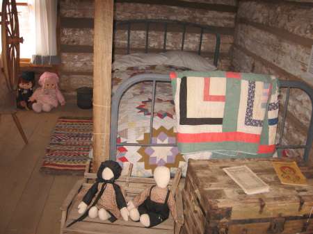 A rustic bedroom with a metal-frame bed, patchwork quilt, wooden chest, dolls on a crate, and woven rugs on a wooden floor.