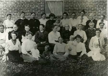 A group of women and children pose outdoors in front of a brick wall for a black and white photograph.