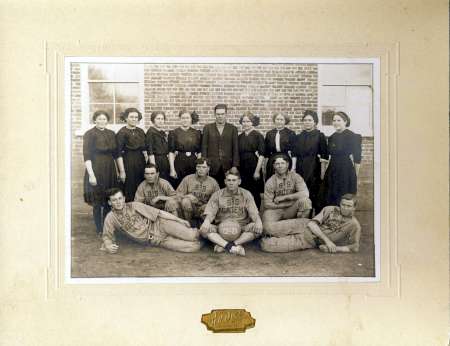 A vintage black-and-white photo of a women's group standing behind a seated men's basketball team in uniforms, posed outside a brick building.