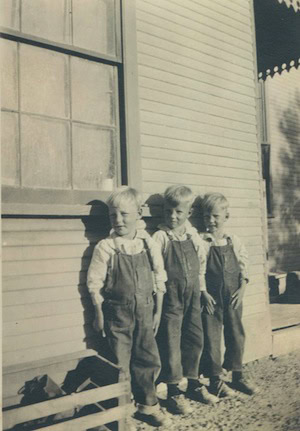 Three young boys in overalls stand side by side outside a wooden building in sunlight, with a window and part of a porch visible.