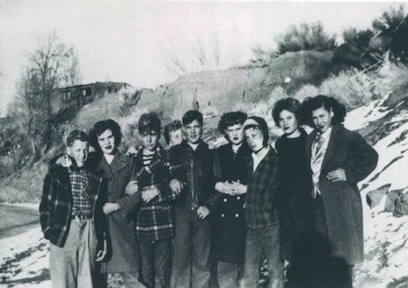 A group of eight people, dressed in winter clothing, stand closely together outdoors in front of a snowy, hilly landscape.