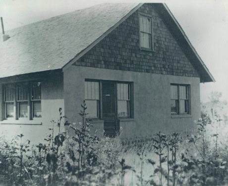 Black and white photo of a small house with a steep roof, shingle siding on the upper section, and overgrown plants in the foreground.