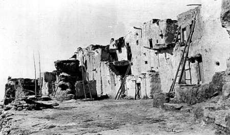 Black and white photo of adobe dwellings with wooden ladders propped against the walls, set on a rocky terrain under a clear sky.