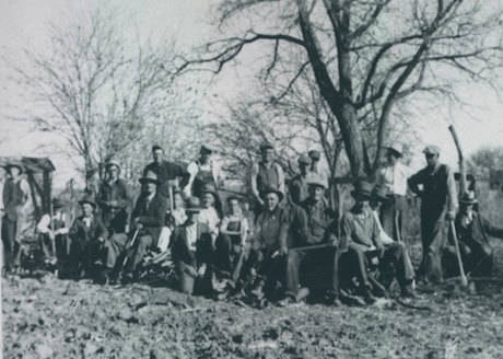A group of people poses outdoors in a rural setting with leafless trees and farm tools, suggesting a farming or work gathering.