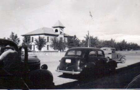 Black and white photo of vintage cars parked on a street in front of a two-story building with a tower and trees.