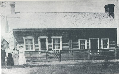 A black and white photo of a wooden log cabin with five windows and a fence in front; four people stand near the left side of the building.
