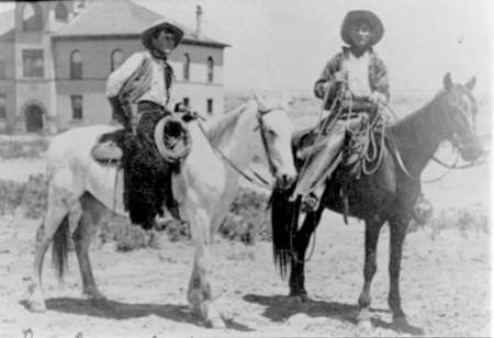 Two cowboys wearing hats sit on horses, holding lassos, with a large building in the background, in a black and white historical photograph.