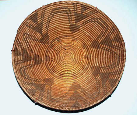 A round, woven basket with a spiral pattern and darker geometric designs radiating from the center. The basket has a natural brown color with subtle variations.
