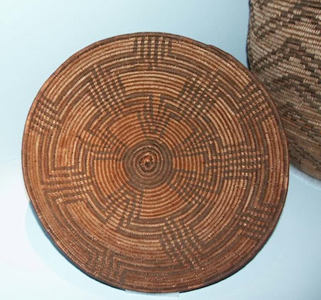 A round, woven basket lid with a detailed geometric pattern made from natural and dark fibers, displayed against a light background.
