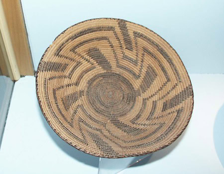 A round woven basket with a geometric black and brown pattern is displayed on a stand against a plain background.