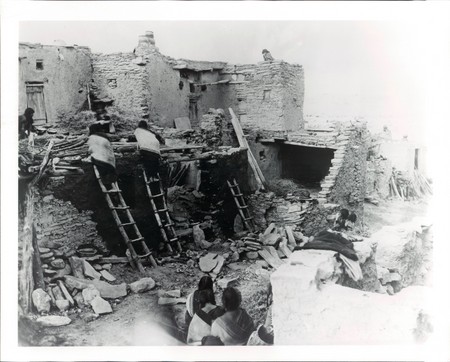 A group of people use wooden ladders to access the roof of a multi-story adobe structure in a historic village setting, with others seated and observing in the foreground.