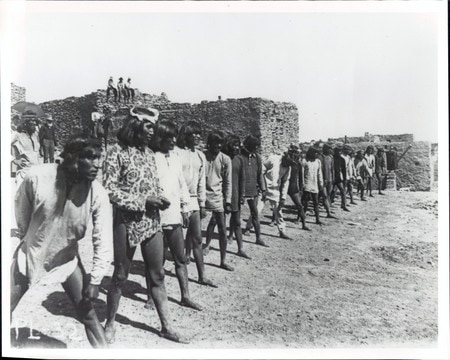 A line of Indigenous men stands in front of a stone building in an outdoor setting, with several people sitting on the structure in the background.