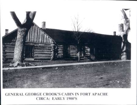 Black-and-white photo of a log cabin with two bare trees in front, labeled as General George Crook’s cabin in Fort Apache, circa early 1900s.