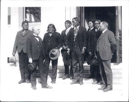 A group of men, including Native American leaders and officials, stand on steps outside a building, dressed in formal suits and holding hats.