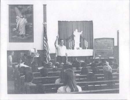 A man stands at the front of a church classroom, raising his hand while children seated in pews raise their hands; an American flag and religious images are visible.