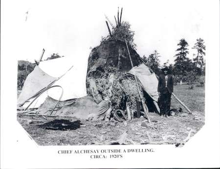 A man identified as Chief Alchesay stands outside a traditional dwelling made of branches and covered with brush and cloth, circa 1920s.