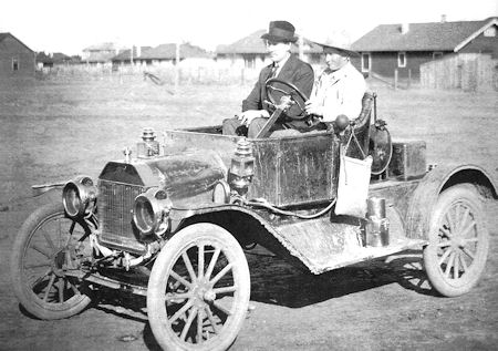 Two people sit in an early 20th-century open-top car on a dirt road with houses and wooden fences in the background.