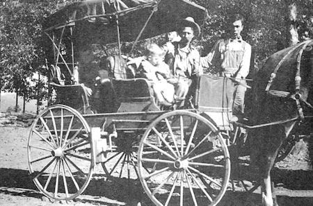 Black and white photo of a horse-drawn carriage with two adults and a child seated inside, and one standing man nearby, outdoors in a rural setting.