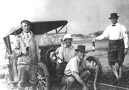Four men in early 20th-century attire pose next to a muddy vintage car; two are kneeling by a wheel, and two are standing, holding bottles.