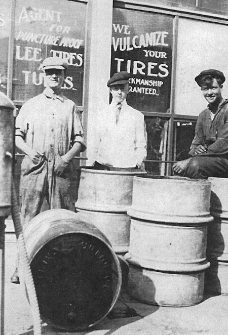 Three people stand outside a tire repair shop with large metal barrels and a tire. Shop windows display signs advertising tire and tube vulcanization services.
