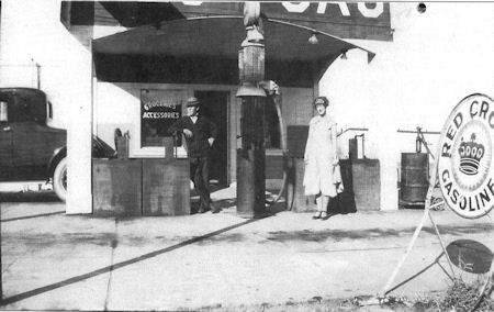 Black and white photo of an old gas station with a man behind a counter and a woman standing near a gas pump; vintage car and Red Crown Gasoline sign visible.