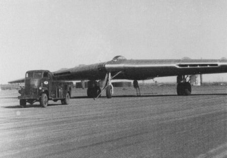 A large aircraft with a distinctive flat wing design is parked on a runway next to a truck; two people are standing near the plane's landing gear.