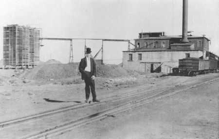 A man in a suit and hat stands near industrial buildings, train tracks, and piles of dirt at an early 20th-century factory site.