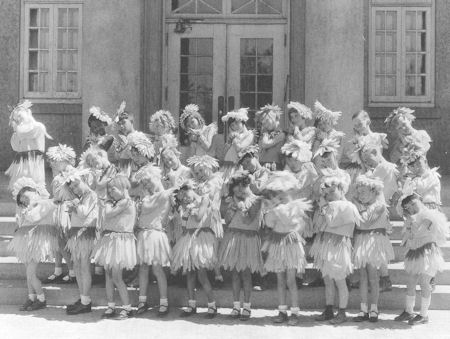 A group of children in grass skirts and feathered headbands stand on steps in front of a building, posing with their arms crossed over their chests.
