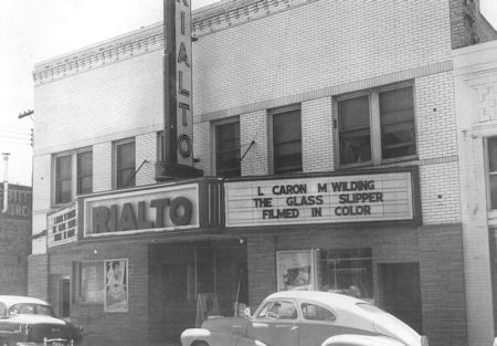 Black-and-white photo of the Rialto Theater with a marquee advertising "The Glass Slipper" starring L. Caron and M. Wilding. Vintage cars are parked in front of the building.
